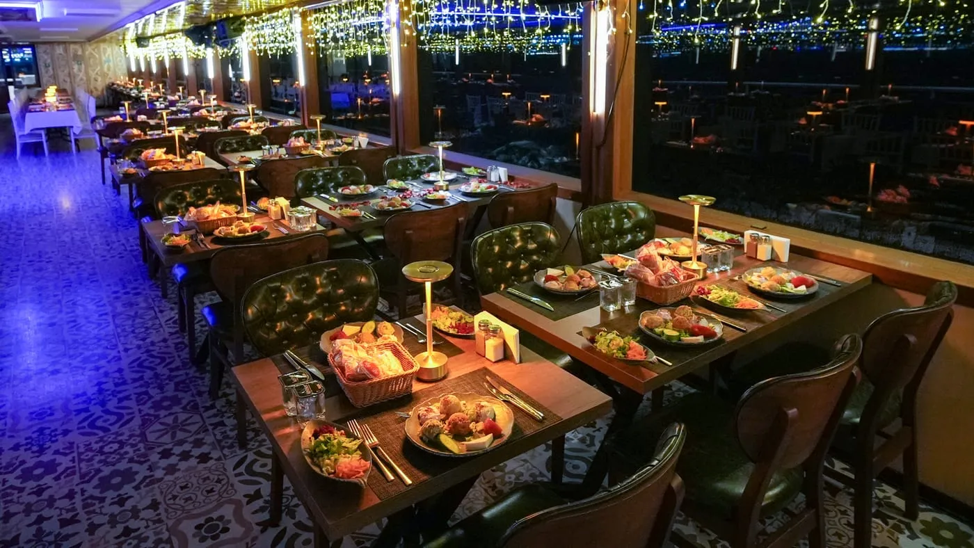 Laid tables in the salon at night, the lit shore beyond the windows