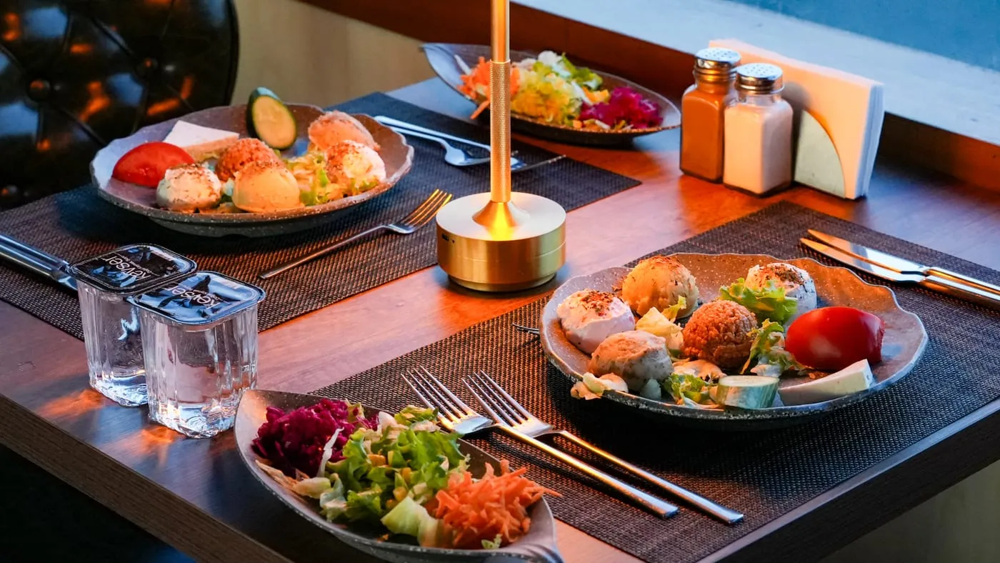 Cold meze plate and salad at a window table, lit by a table lamp