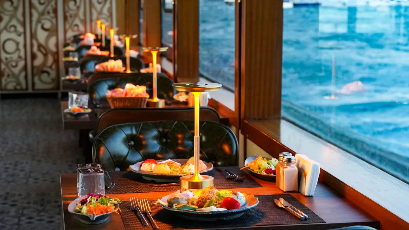 A row of window tables with lamps lit and the water beside them
